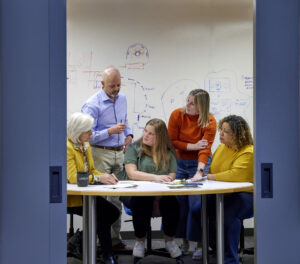 Peering between two open sliding blue doors , five team members are huddled around a table talking to one another in front of a white board