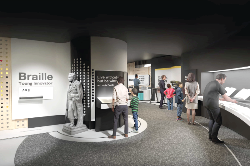 A wide shot of the exhibits. Toward the left, is a statue of Louis Braille. To the left of the statue, on the wall, says, “Braille. Young Inventor.”